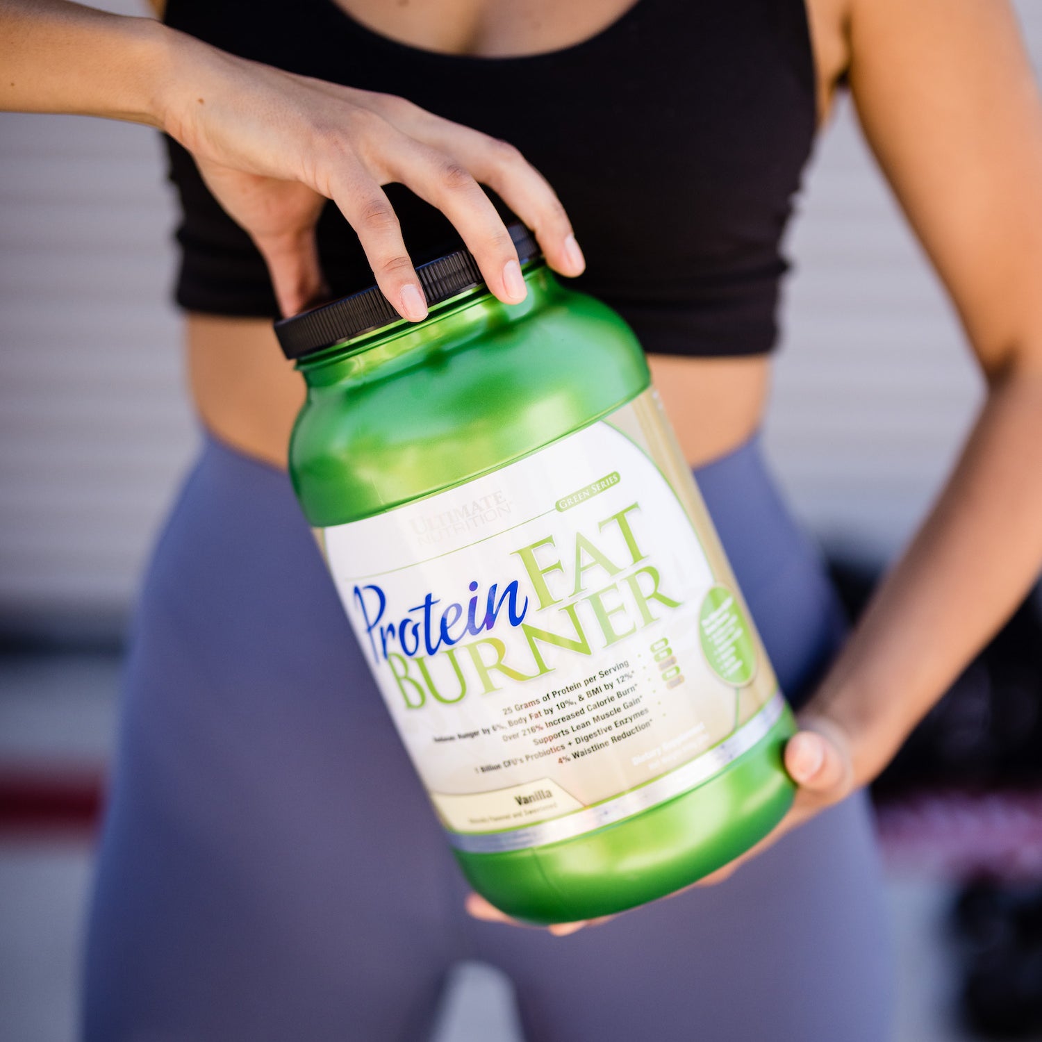 An athlete in workout attire holds a green container of Protein Fat Burner supplement, showing the product label against a blurred background