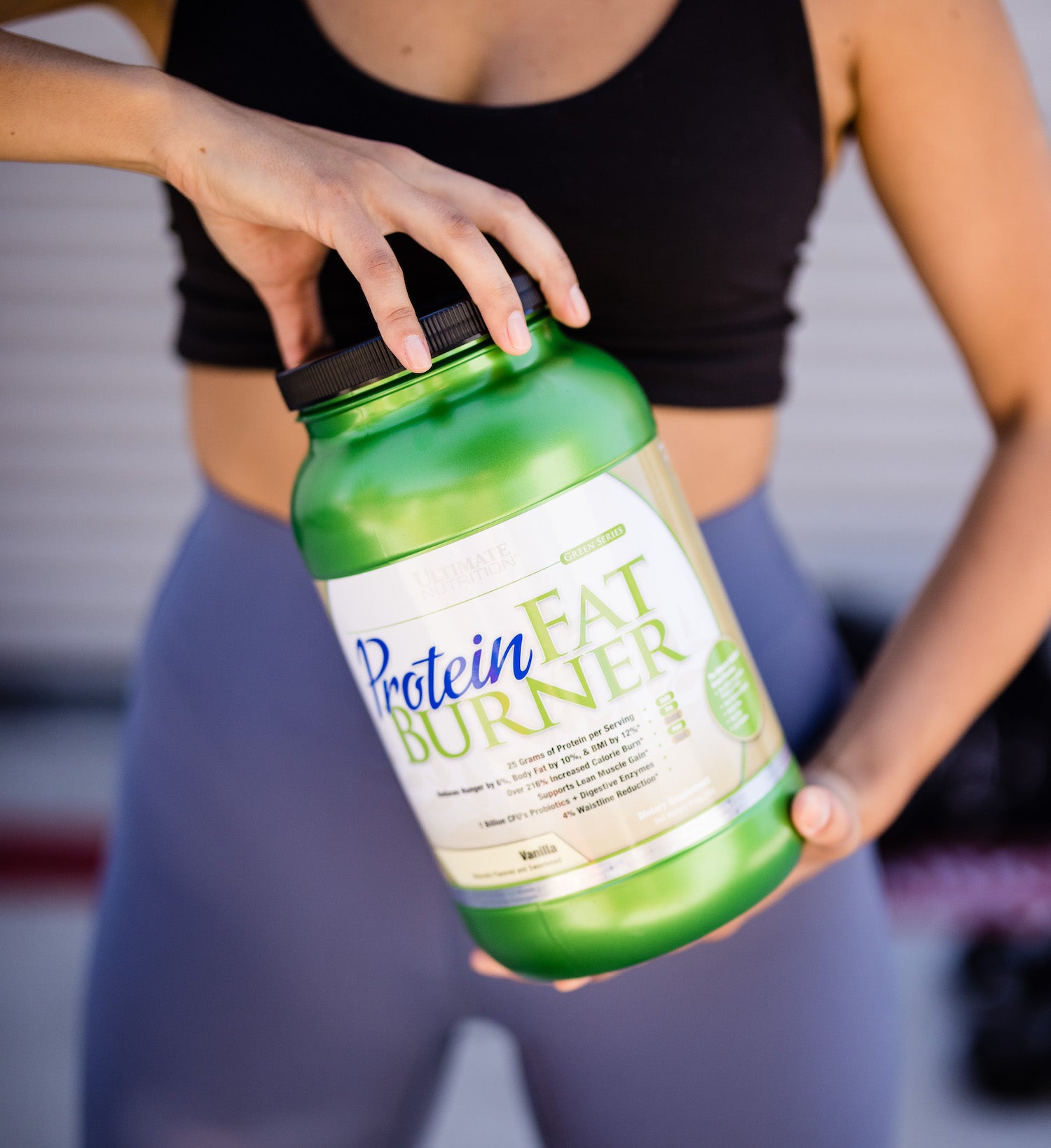 An athlete in workout attire holds a green container of Protein Fat Burner supplement, showing the product label against a blurred background